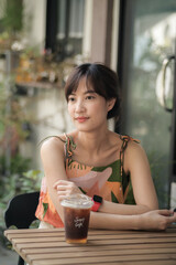 Asian woman in tropical patterned dress sits at outdoor cafe table with cup of iced coffee, enjoying serene moment