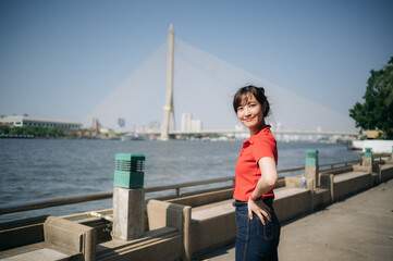 Asian cheerful woman in red shirt poses confidently by riverside with cable stayed bridge in background under clear blue sky