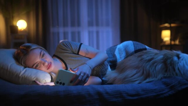 Young woman scrolling on her smartphone in bed at night, yawning, and falling asleep with her dog beside her. Cozy evening routine