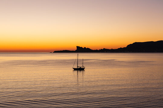 Coucher de soleil sur la mer en M&eacute;diterran&eacute;e avec un voilier et rochers en arri&egrave;re plan sur fond de ciel orang&eacute;