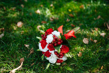 Obraz premium A vibrant wedding bouquet with red roses, white flowers, greenery, and red berries resting on green grass outdoors. 