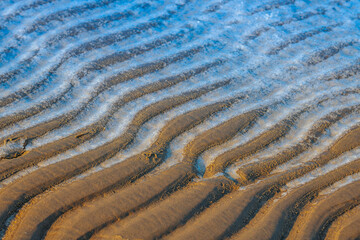 Close-up of wet sand ripples with dark and light patterns formed by receding water, creating a textured and natural abstract design. A mix of golden, brown, and blue tones enhances the contrast.