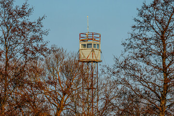 A rusty old watchtower with a weathered metal frame stands against a clear blue sky, with bare tree branches in the foreground, creating a nostalgic and abandoned atmosphere.