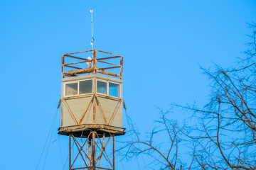 A rusty old watchtower with a weathered metal frame stands against a clear blue sky, with bare tree...
