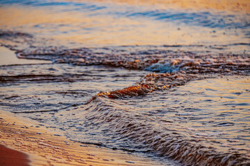 Gentle ocean waves roll onto the sandy shore, illuminated by warm golden sunlight. The shimmering water reflects the hues of the sky, creating a tranquil and scenic coastal view.