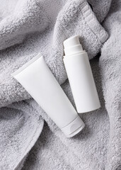 White blank cream tube and bottle on light grey bath towel top view, cosmetic mockup