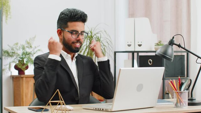 Indian businessman freelancer taking break leaning on chair after working celebrating success victory at modern home office workplace desk. Male Hindu in formal suit puts hands behind head relaxing