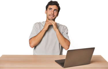 Hispanic young man working on laptop keeps hands under chin, is looking happily aside.