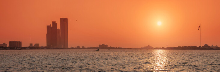 Scenic view of Abu Dhabi skyline reflecting on the sea at sunset with United Arab Emirates flag waving