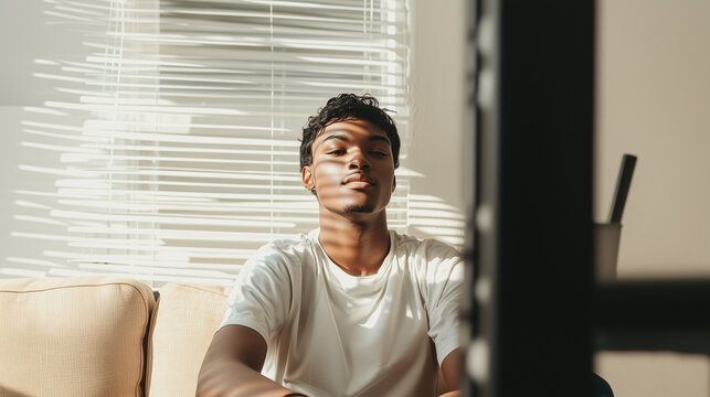 A young man sits on a sofa with shadows from window blinds cast across his face. A quiet, introspective moment filled with aesthetics of light and shadow. - Powered by Adobe