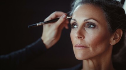 Obraz premium Makeup Artist Applies Cosmetic Brush to Mature Woman s Face in Beauty Portrait against Dark Backdrop