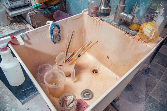 Close-up of a sink in a pottery studio filled with assorted pots, paintbrushes and cleaning materials