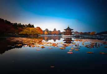 Fototapeta premium Serene Chinese landscape featuring a pagoda reflecting in calm water. Autumn foliage adorns trees around the tranquil lake under a clear blue sky.
