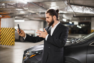 Young Caucasian male in suit speaking on phone video call in underground car park next to car. Business communication concept with focus on technology and modern work style.