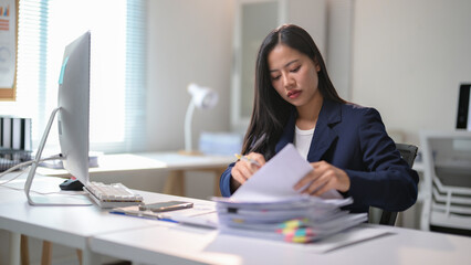 A woman is sitting at a desk with a computer monitor and a stack of papers