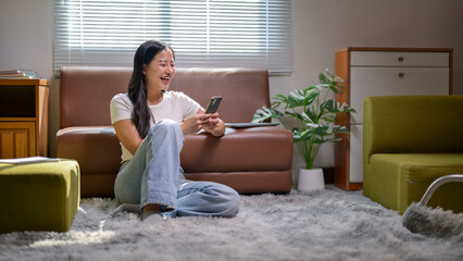 A woman is sitting on the floor in a living room, looking at her cell phone