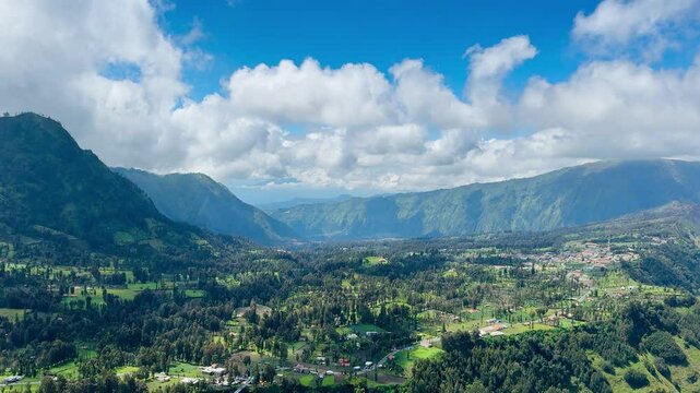 Aerial view of a green village from Sunrise Point Bromo, Sukapura, Probolinggo, Indonesia. Lush valley, misty hills, and floating clouds create a serene Swiss-like highland landscape.