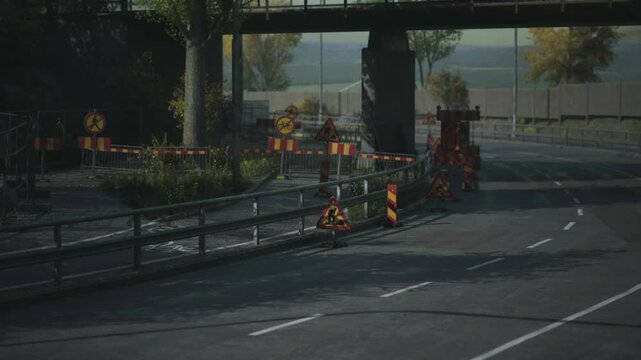 Workers are engaged in construction activities along a roadside under a bridge. Traffic signs indicate detours and safety measures during the autumn season.