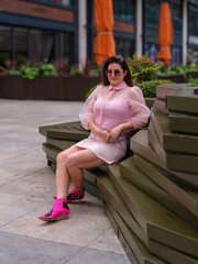 A woman in a pink dress with sheer sleeves and bright pink boots sits confidently on a modern...