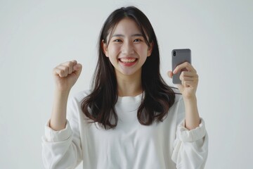 Fototapeta premium Elated Asian woman holding her smartphone, celebrating her big win with a wide smile, white backdrop