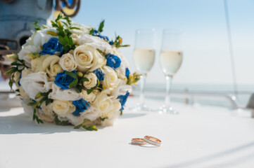 A wedding scene featuring blue and white floral bouquet, wedding rings, and champagne glasses, set against a sunny outdoor backdrop.
