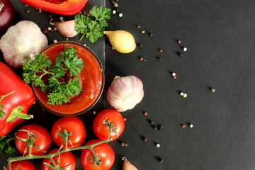 On a black background lies a stone tray with a glass bowl of tomato sauce and fresh vegetables.	