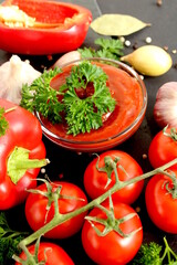 On a black background lies a stone tray with a glass bowl of tomato sauce and fresh vegetables.