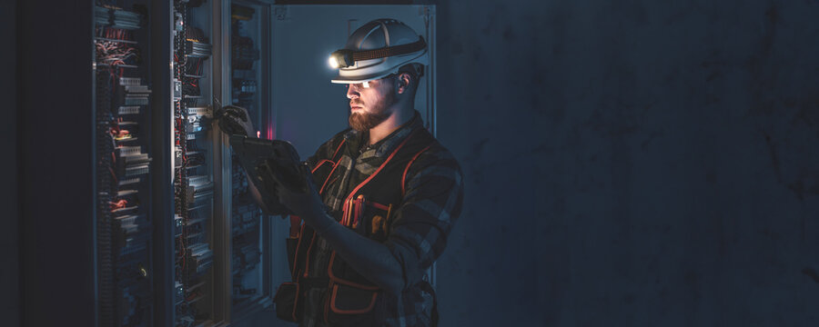 A male electrician in overalls, focused on work in switchboard with fuses, using tablet.