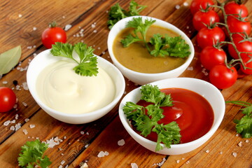 There are three white bowls with mayonnaise, ketchup and mustard on a wooden table.	