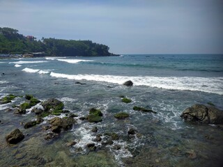 A beach with gentle waves, mossy rocks, and green hills in the distance. Fishing boats line the shore, creating a serene and natural coastal atmosphere.