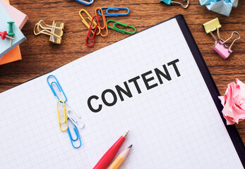 The word CONTENT written on a white grid notebook on a wooden table. There are various office supplies such as paper clips, sticky notes, pencils, and crumpled paper nearby.