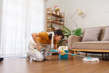 Asian housemaid scrubbing the wooden floor in a modern living room with cleaning supplies