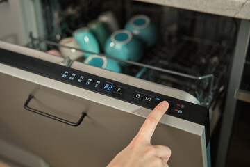 Close-up of female finger pressing button on dishwasher control panel. Woman using dishwasher in...