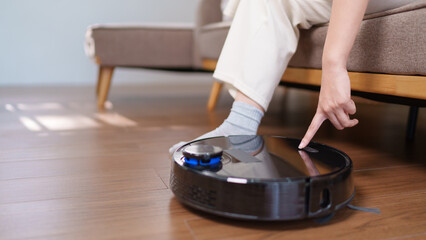 Asian woman turning on a robotic vacuum while sitting on a sofa in a cozy living room