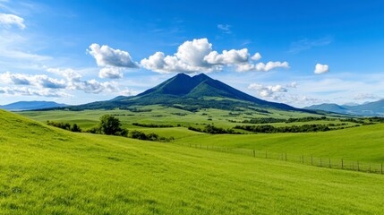 Panoramic view of a lush green valley and a volcanic peak beneath a vibrant blue sky speckled with fluffy white clouds. The foreground displays
