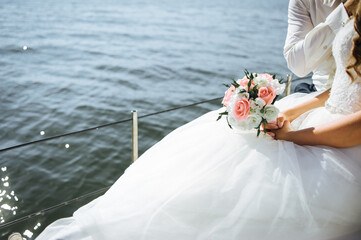 A bride in a white dress holding a floral bouquet while sitting on a yacht, overlooking calm blue...