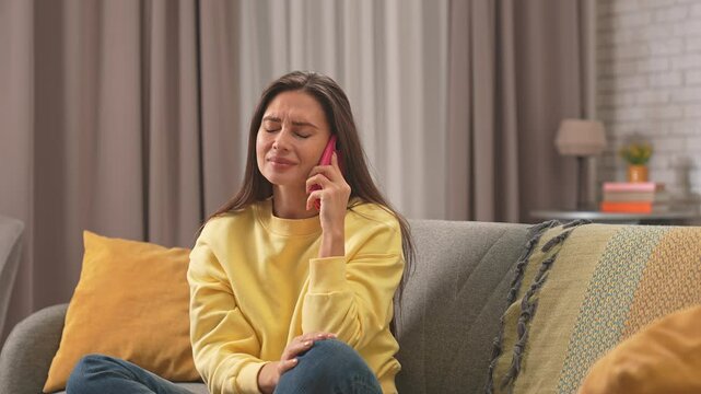 Young woman spending day at home sitting on the sofa in the living room, talking answering call on smartphone, upset apologizing expression.