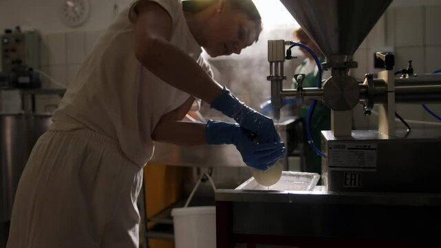 A female cheese maker fills burrata using special equipment, forms a cheese ball and ties it. Production of burrata and mozzarella in a cheese factory, industrial space, food.