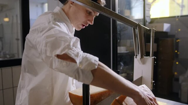 A woman in white cuts hard cheeses with a large industrial knife into small pieces to display on the counter. Slicing cheese for sale at a cheese factory.