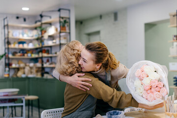 A young Caucasian woman surprises her elderly mother with a floral bouquet at a creative workshop. They celebrate a special event together, expressing love and joy in a cozy florist studio.
