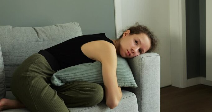 Discouraged female teenager hugs cushion lying on sofa and opens sad eyes. Girl experiences negative emotions due to misunderstanding with friends