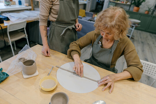 Teacher guides an elderly women, demonstrating sculpting techniques in a hands-on class. Elderly Caucasian woman learns pottery at a creative workshop with a young mentor