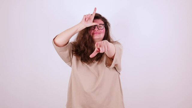Brunette young plus size woman in beige t-shirt making loser gesture, L sign on forehead, teasing, accusing for defeat, expressing disrespect. Indoor studio shot isolated on white background. you lose