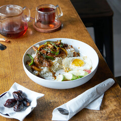 A flavorful dish featuring rice, green peppers, tender meat, and a sunny-side-up egg sits on a wooden table alongside tea and dates