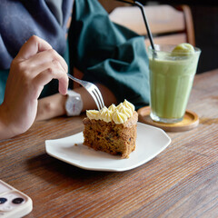 A person is savoring a slice of moist banana cake topped with frosting, paired with a chilled green drink in a warm cafe atmosphere