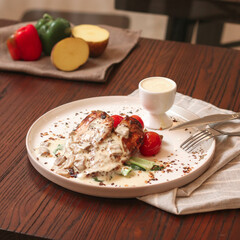 Grilled chicken is topped with a creamy sauce, served with tomatoes and vegetables on a modern plate in a warm dining environment