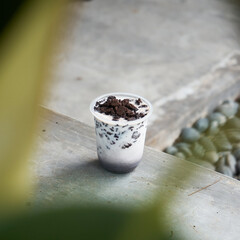 A decadent dessert cup filled with creamy layers topped with crushed cookies sits on a stone table amidst green plants