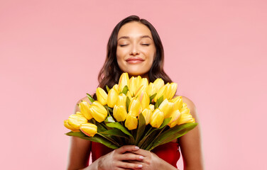 Happy lady with closed eyes sniffing bouquet of yellow tulips, enjoying aroma on pink background. Romantic gift, congratulation at women's day