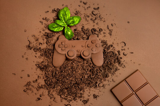 A chocolate game controller with detailed craftsmanship lies on a bed of chocolate shavings, complemented by a fresh green basil leaf and a chocolate bar, blending gaming and indulgence creatively.