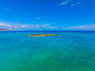 Lush green island surrounded by turquoise waters, with small boats and paddleboarders nearby, captured in an aerial view near Oahu, Hawaii.